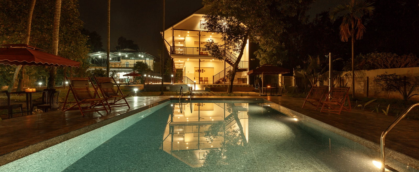 A swimming pool illuminated at night, with buildings and trees visible in the background.