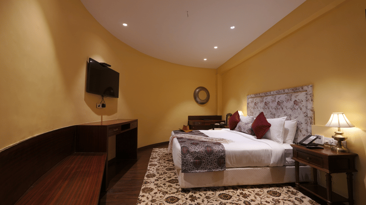 Side view of the double bed, a wall mounted television and a wide table in the Suite Cottage Room at The Garden Ananta Elite Rajkot 4