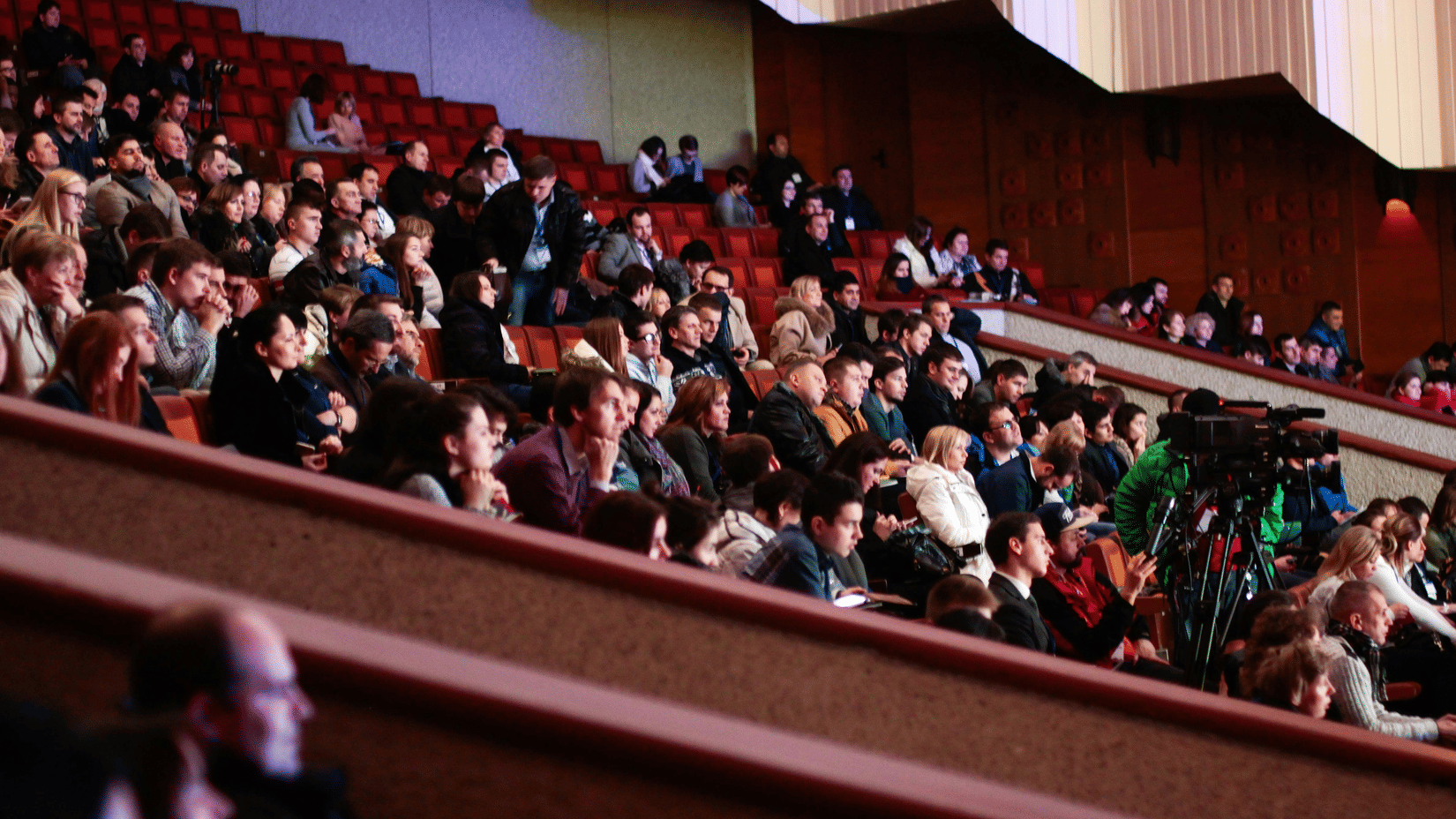 A large crowd of people seated in a tiered auditorium or theatre, viewed from a side angle during an event.