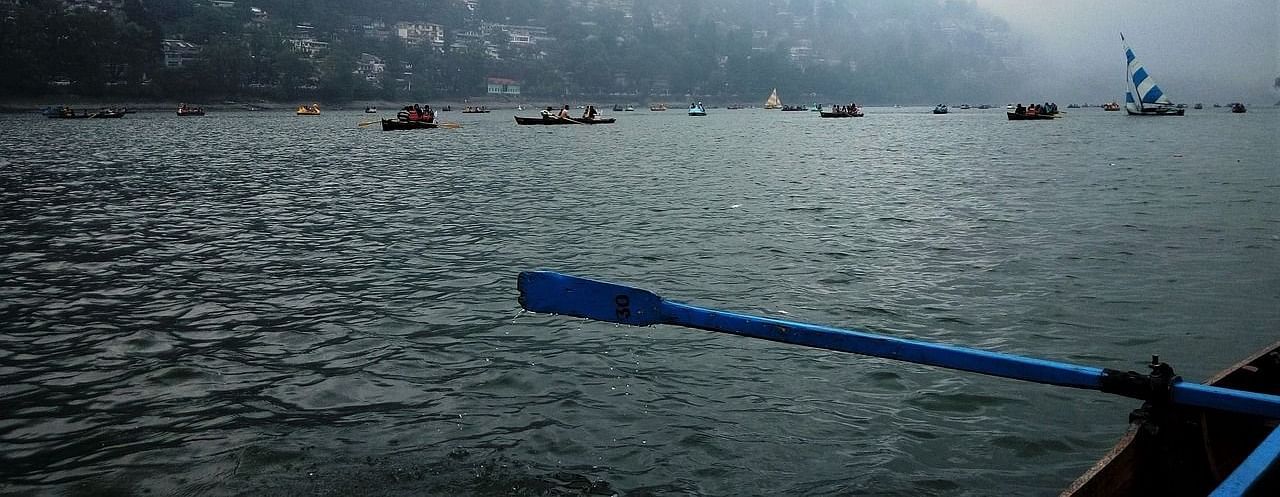 A scenic view from a raft on Nainital Lake, featuring misty hills in the distance and fellow boaters gliding across the shimmering blue water, evoking a sense of tranquility and adventure .