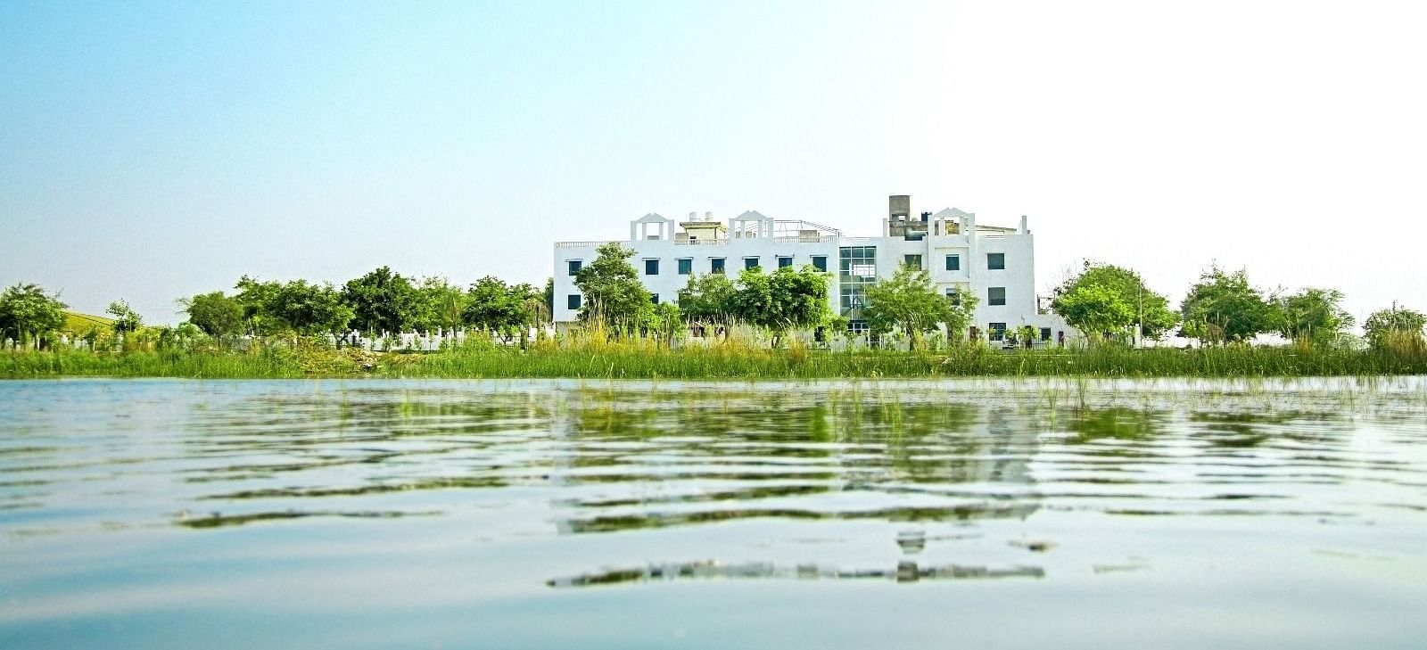 A lake view, with Grassfield Riveria Resort, Jaipur, visible on the shore surrounded by greenery under a clear sky.
