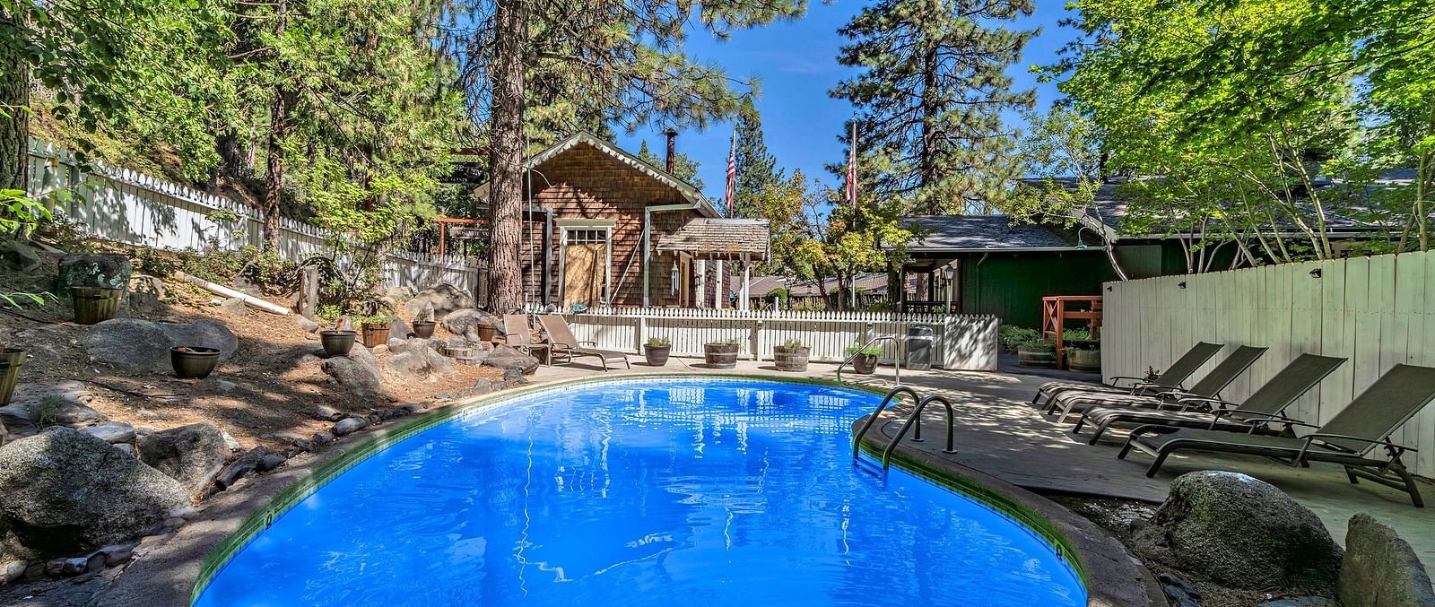 A view of the outdoor swimming pool with a cabin in the backdrop surrounded by trees on a clear blue day