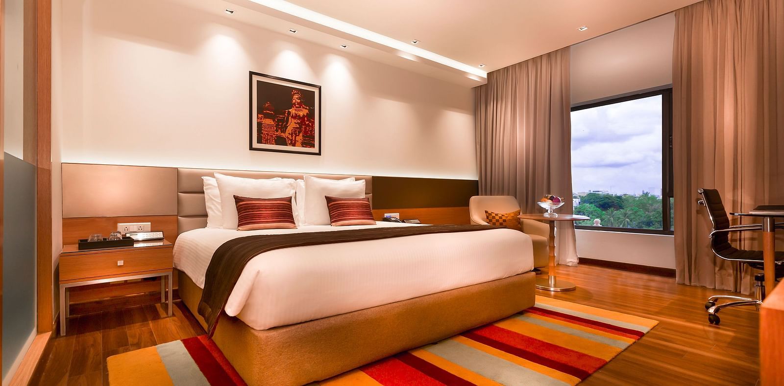 A modern hotel room featuring a king size bed, warm lighting, wooden flooring, a striped rug, and a large window with a view - Grand Chennai by GRT Hotels