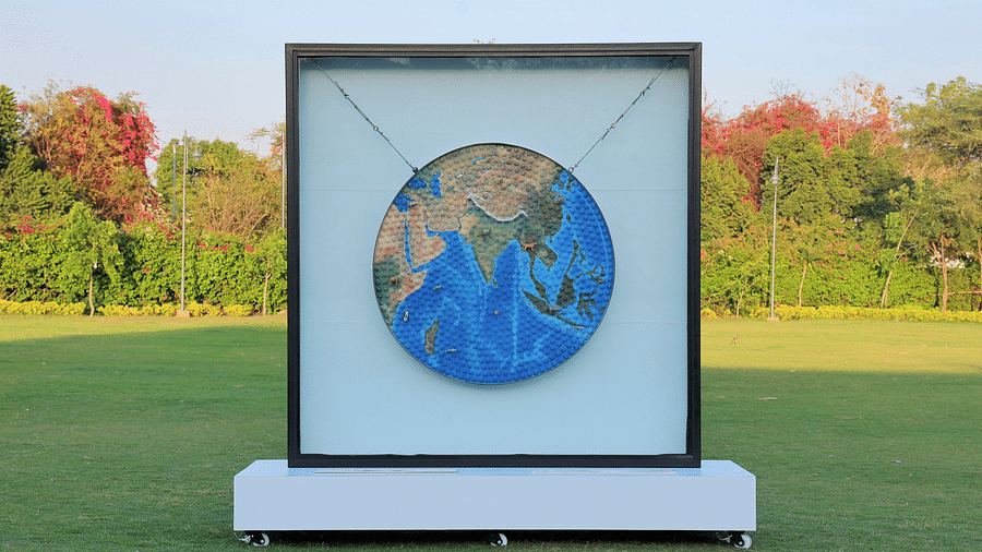 A circular 3D map art installation depicting Asia and the Indian Ocean, mounted on a stand in an outdoor setting with greenery and trees in the background - Karma Lakelands.