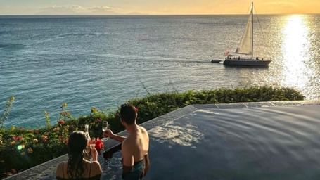 A couple enjoying drinks in an infinity pool overlooking the ocean at sunset, with a sailboat gliding across the water in the distance.