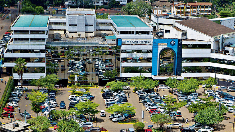 Aerial view of the large commercial building of Sarit Centre in Nairobi with glass windows and an entrance archway overlooking a busy parking area filled with many vehicles.