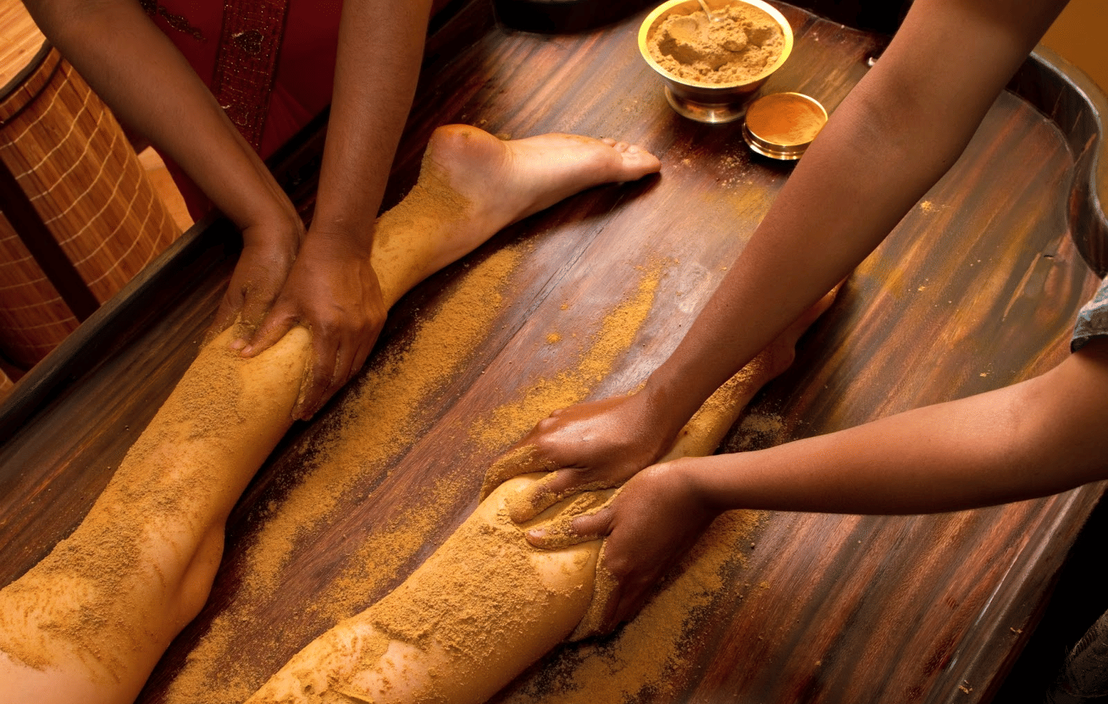 Close-up of hands applying herbal paste on a person's legs during a massage, with a wooden surface and bowl of paste visible at Paradise Lagoon Resort, Udupi.