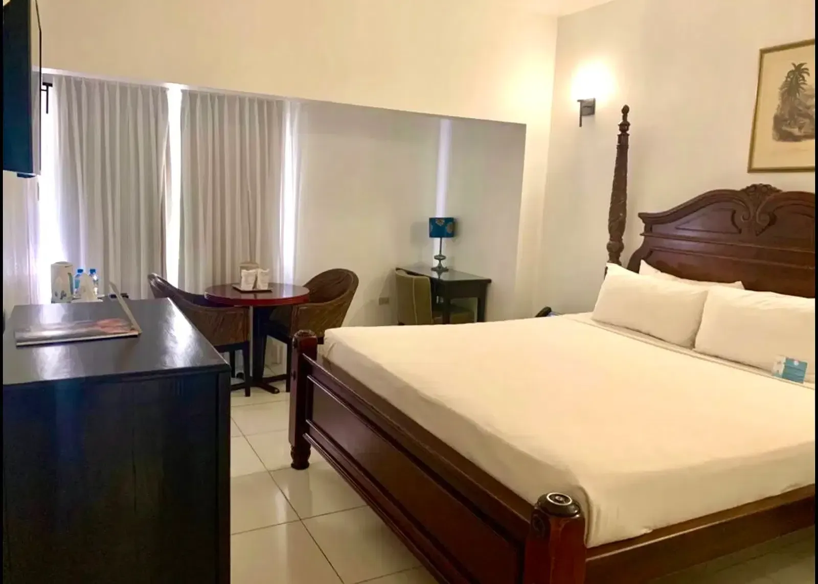 A well-lit hotel room features a large wooden bed with white linens, a dark dresser, and a seating area near a window | Executive King Room | Altamont Court, Jamaica