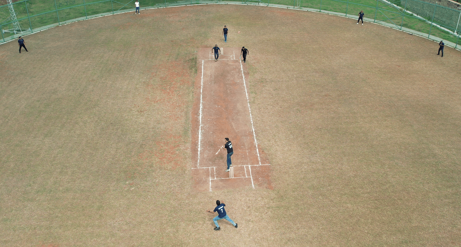 Aerial view of a cricket match in progress at Ramoji Film City, showing players positioned on the field with a bowler mid-action and the batsman ready to strike.