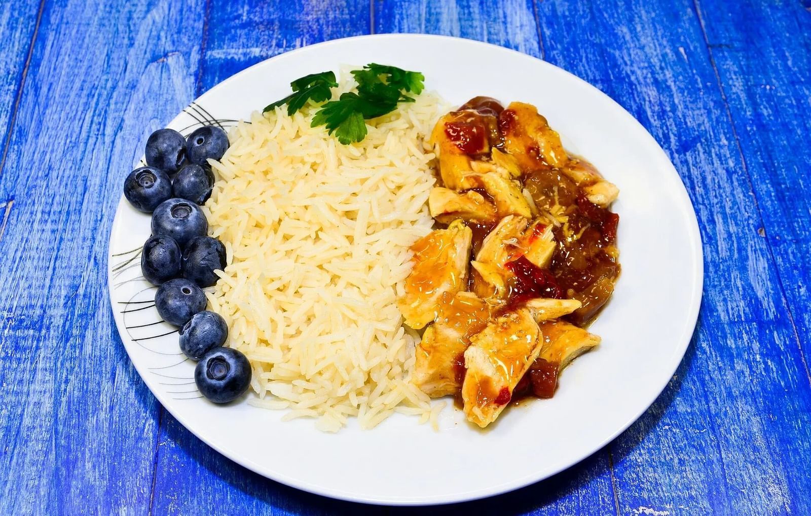 A white plate holds a serving of rice, with meat, and blueberries, presented on a blue textured surface | Altamont Court, Jamaica