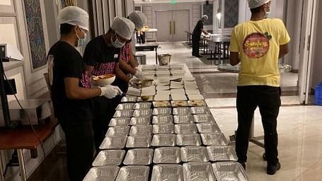 The staff filling up a lot of aluminium  foil containers with food wearing masks and gloves.