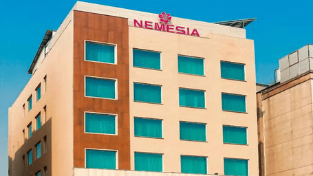 Daytime view of the Nemesia hotel building facade with teal-tinted windows, wooden accents, and palm trees in front.