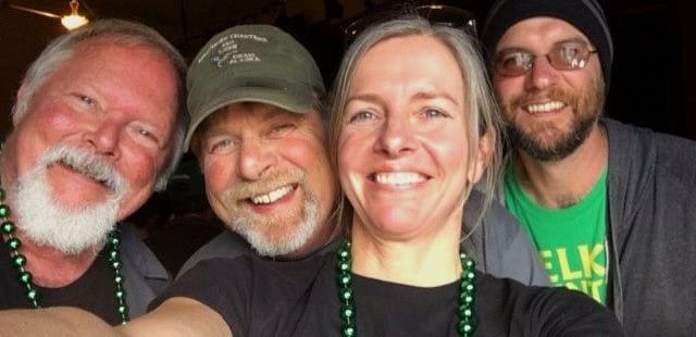 A group of four smiling friends wearing green beads, enjoying a night out at a bar or brewery