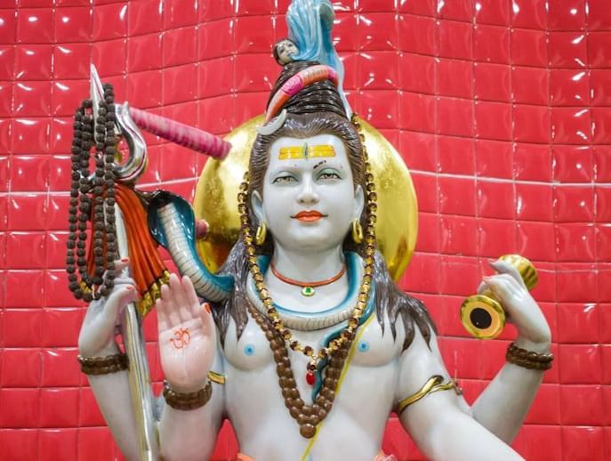 Colourful statue of a Hindu deity holding a trident and a drum placed against a red background.