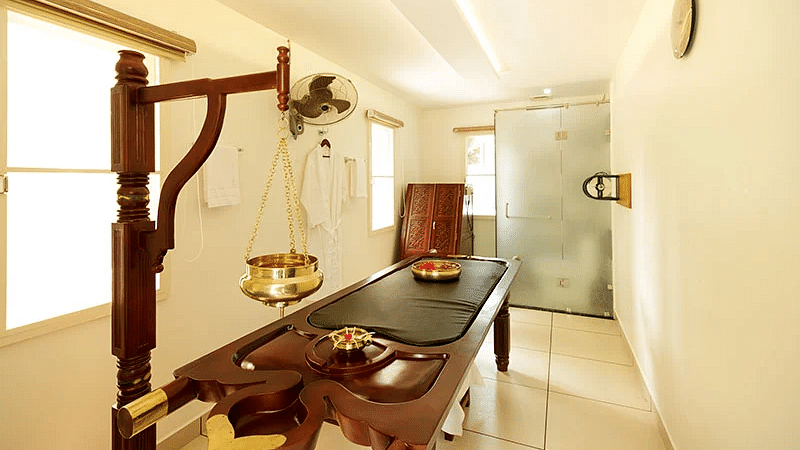 A traditional wooden massage table in a white room with natural light, suggesting a treatment area at Lhasa Ayurveda & Wellness Resort.