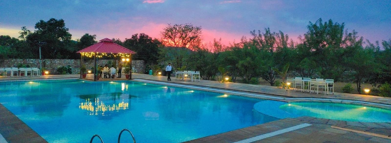 An outdoor swimming pool at dusk with a pink and blue sky in the background - Bamboo Saa Mulberry Resort, Pushkar