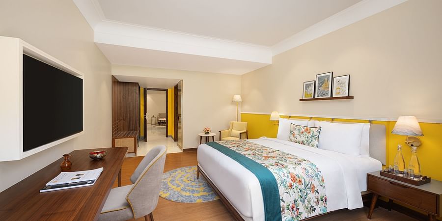 A well-lit hotel room with a queen bed featuring floral and teal accents, a wooden desk with a chair and a television, a nightstand with a lamp, and an open doorway leading to another room - Aloha On the Ganges.