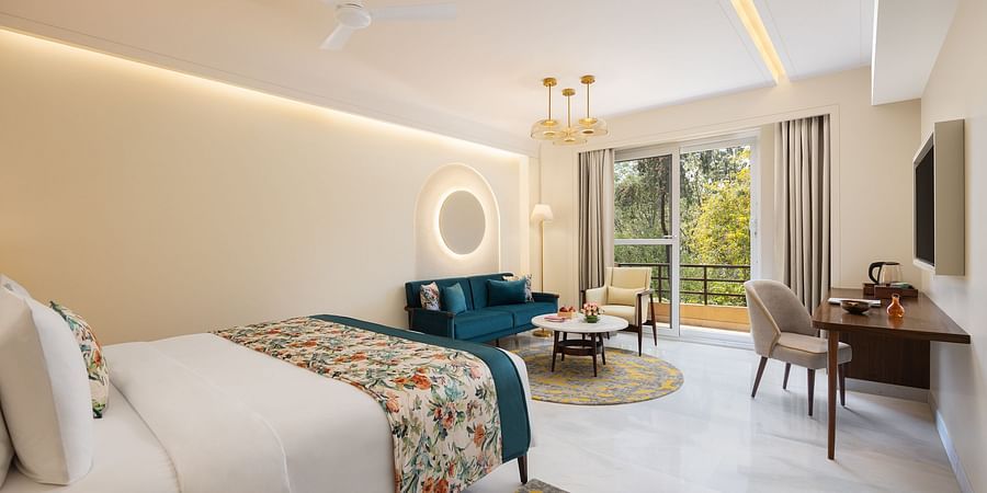A bright and airy hotel room featuring a queen bed with floral accents, a teal velvet sofa, a round coffee table on a patterned rug, a work desk with a chair and a television, a balcony with outdoor seating and a view of greenery, modern lighting, and a c