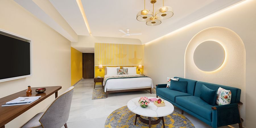 A modern hotel room with a queen bed featuring floral accents, a blue velvet sofa with decorative pillows, a white round coffee table on a yellow and grey rug, a wooden desk with a chair and a wall-mounted television, stylish lighting fixtures, and yellow