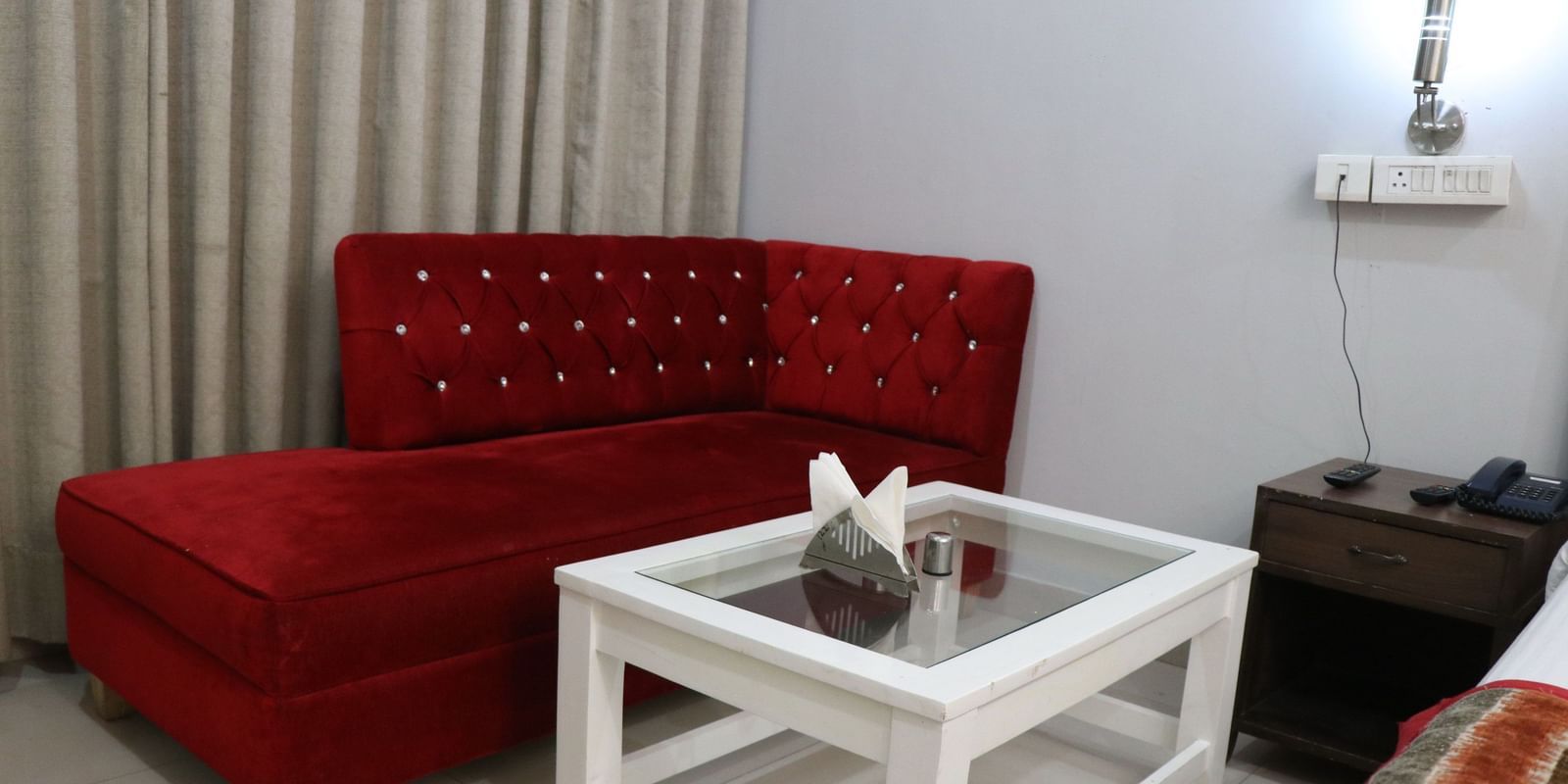A cosy corner with a red tufted sofa, a white square table, and a bedside table, illuminated by a wall lamp against a soft gray wall. - Hotel Diamond Plaza, Chandigarh.