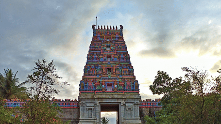 A wide stone pathway leading directly toward a tall, intricately designed Hindu temple tower surrounded by green trees.