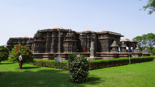 The back view of Hoysaleswara Temple, Halebidu, having intricate stone carvings on it, a lush garden in the foreground and blue sky in the background.