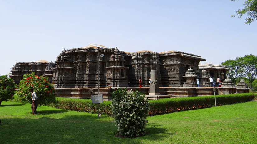 The back view of Hoysaleswara Temple, Halebidu, having intricate stone carvings on it, a lush garden in the foreground and blue sky in the background.