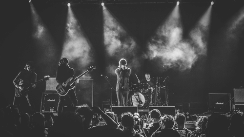 A band performing live on stage with bright lights shining down, a singer with a microphone, and an audience watching the concert
