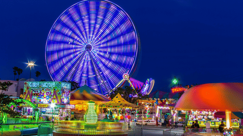A vibrant funfair at night featuring a large, brightly lit Ferris wheel with purple motion-blur lighting effects.