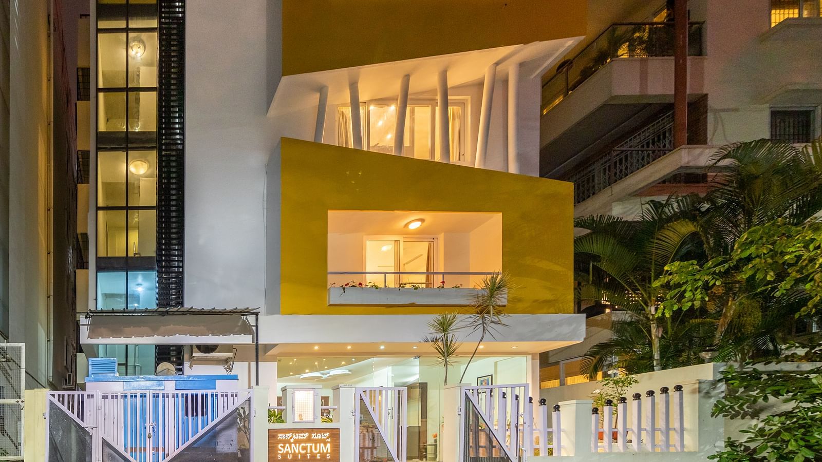 A facade of Sanctum Suites Domlur, Bangalore with warm lighting and view of balconies shot at night.