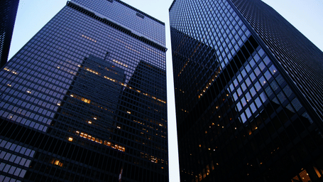 Two tall dark glass skyscrapers photographed from a low angle against a clear bright blue sky, with city buildings visible in the background