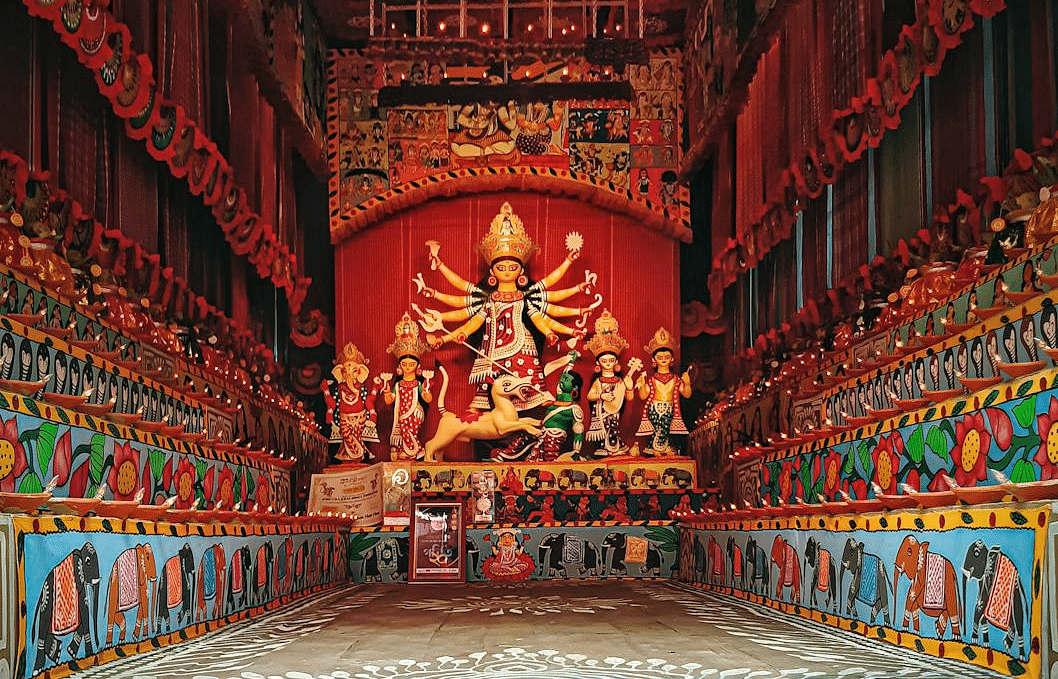 A depiction of Devi Durga surrounded by other gods and goddesses, with an asura being defeated, and small oil lamps lit all around the scene
