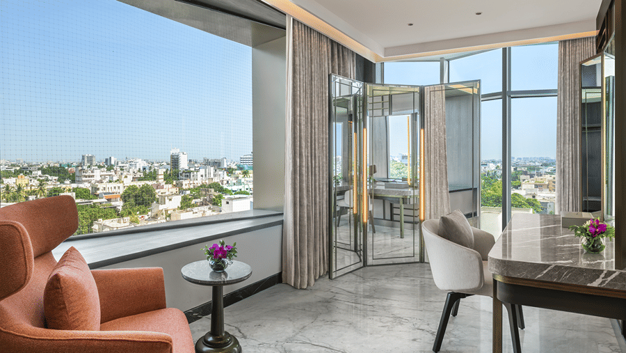 Bride room with an orange armchair by a city-facing window, a marble dressing table with a chair and a mirrored folding screen at The Residency Towers Chennai.