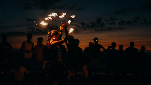 A person performing with fire sticks at night surrounded by a group of people watching in the background.