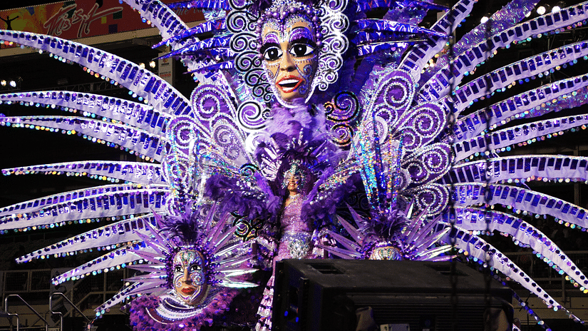 corner view of a carnival decoration in purple colour with large face masks with design on them and a woman dancing on the centre of the stage