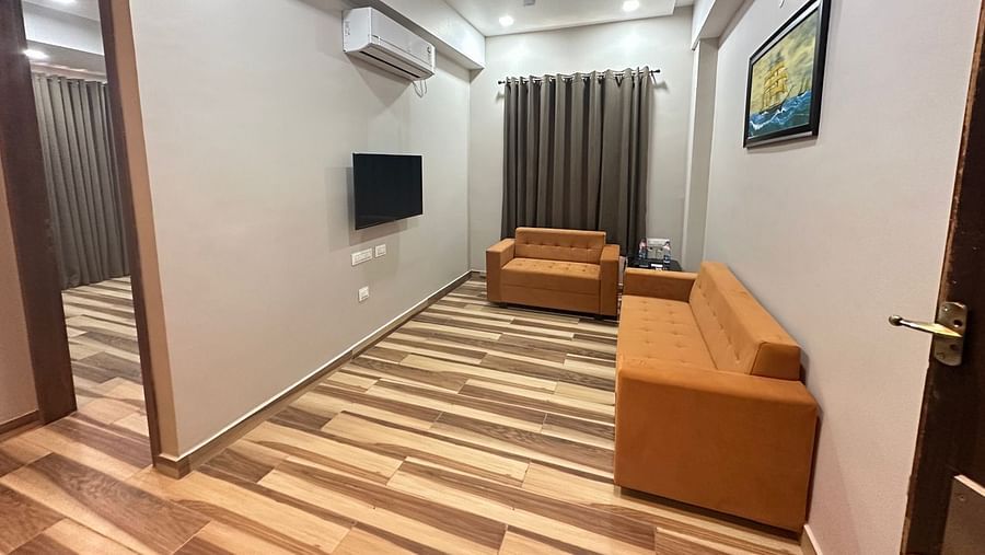 A room features wooden flooring, a brown sofa, a television on the wall, and a ceiling fan - Kankidham Lords Inn