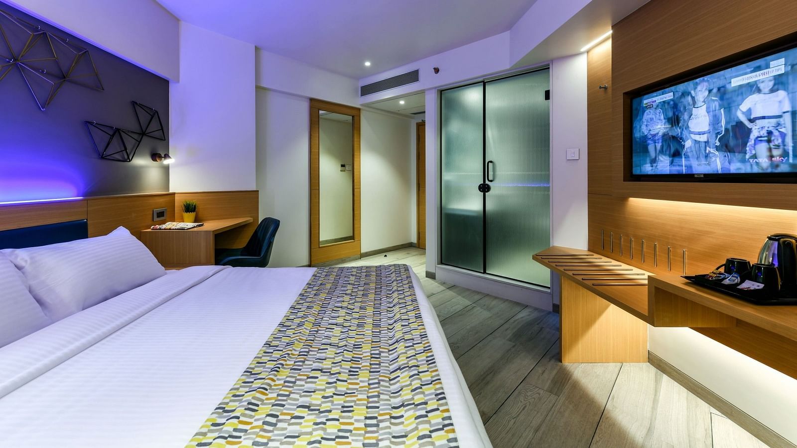 A spacious room with a large bed, a wall-mounted television, and a desk positioned below it, illuminated by cool lighting at Indie Stays, BKC Kalina.