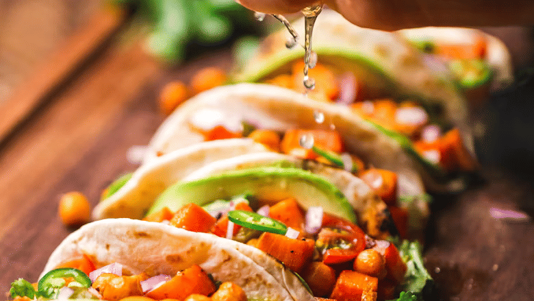Hand squeezing lime juice over a row of soft-shell tacos filled with roasted chickpeas, diced vegetables, avocado, jalapeño, and coriander.