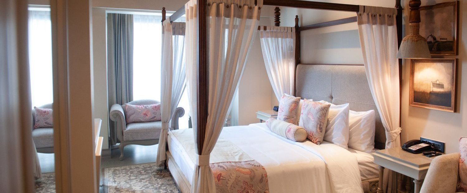  A detailed view of the four-poster bed in an executive room at The Evren, Vagator. The room has a vaulted wooden ceiling, a ceiling fan, and a seating area near the window. The bed is adorned with decorative pillows and curtains.