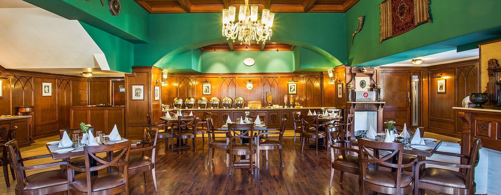An image showing the interior of restaurant at The naini retreat, Nainital. featuring wooden decor, flooring, and a wooden bar. A large chandelier hangs from the ceiling, providing ambient lighting.