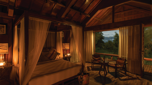A room at The Serai Bandipur lit up at night with a 4 poster bed,  chairs, and large windows