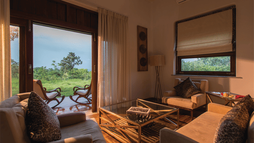 Sun illuminating the interiors of a room at The Serai Bandipur, one of the best Romantic Getaways near Bangalore, featuring a sofa set and a patio seen through the doorway with a couple of chairs facing the trees outside