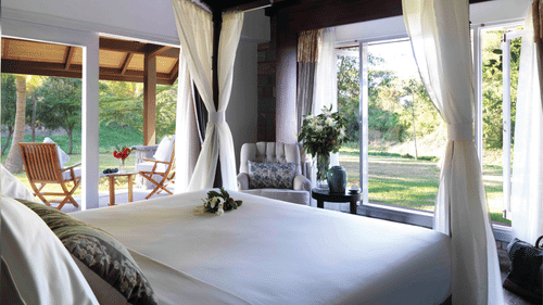A bright, airy bedroom at The Residence, The Serai Kabini, with a canopy bed and sheer curtains, large windows opening to lush greenery, an armchair, and a covered patio with wooden chairs overlooking the garden for a relaxing stay in Kabini.