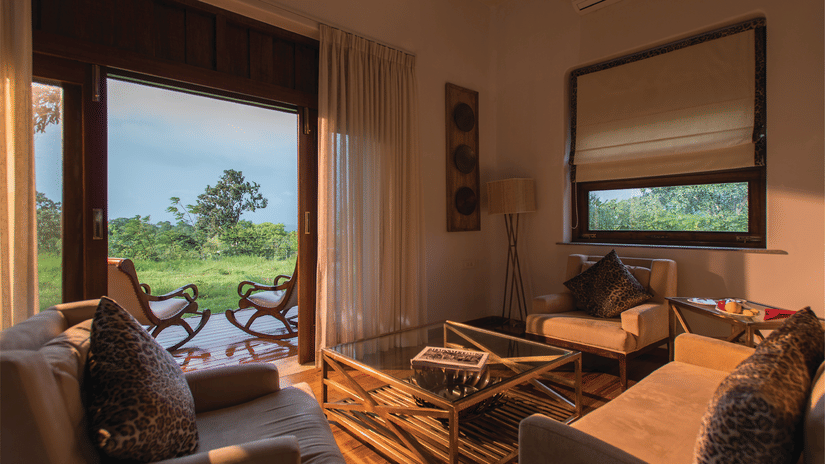 Sun illuminating the interiors of a room at The Serai Bandipur, one of the best Romantic Getaways near Bangalore, featuring a sofa set and a patio seen through the doorway with a couple of chairs facing the trees outside
