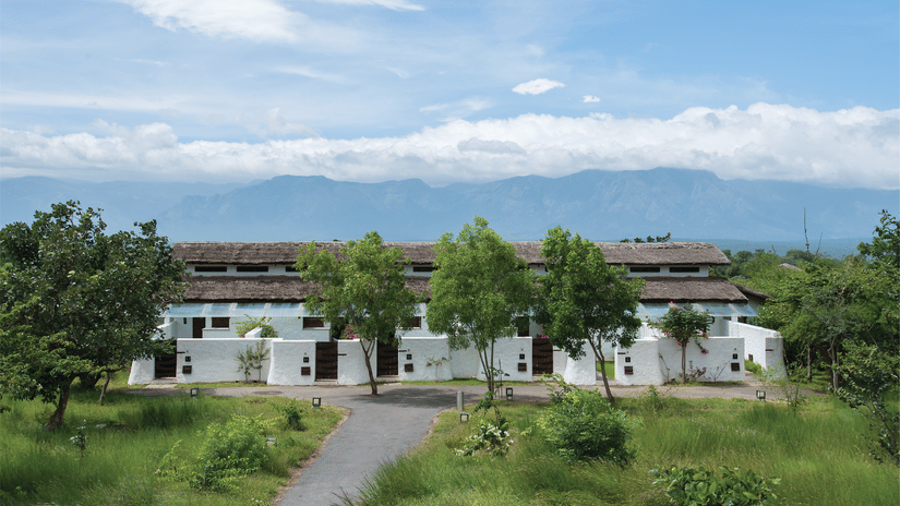 The exterior view of Serai Bandipur, surrounded by trees and greenery, with a pathway leading to the entrance and mountains visible in the distance.