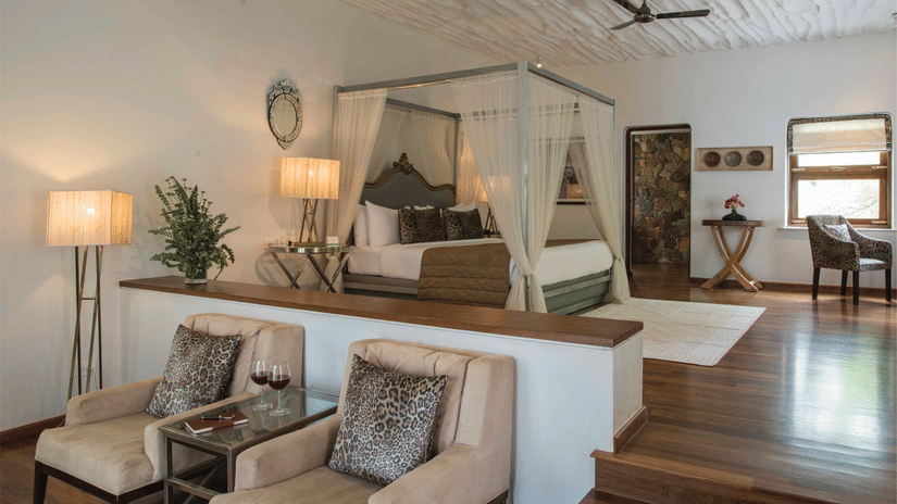 A bedroom at The Serai Bandipur with a 4-poster bed, chairs, side tables, lamps, artwork, and wooden flooring, divided by a small steps.