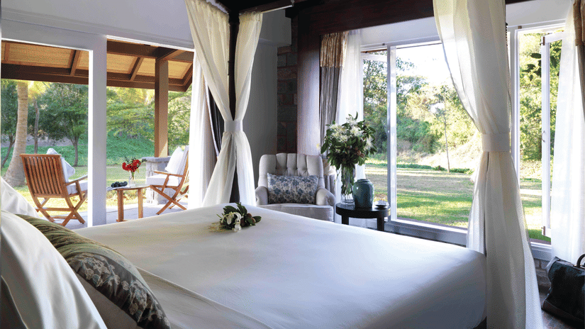 Bright, airy bedroom at The Residence, The Serai Kabini, with a canopy bed and sheer curtains, large windows opening to lush greenery, an armchair, and a covered patio with wooden chairs overlooking the garden for a relaxing stay in Kabini.