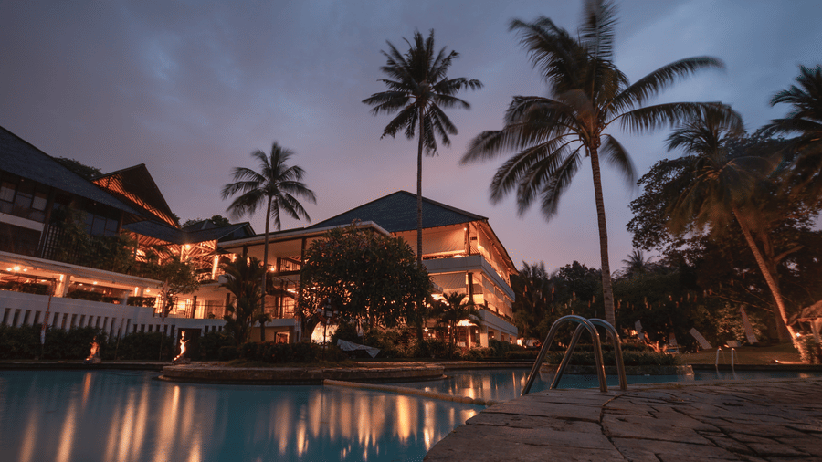 a picture of a Resort next to a swimming pool with coconut trees next to it and captured during sunset