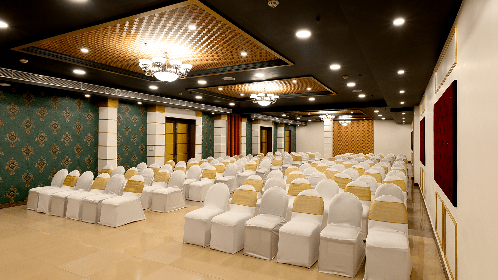 A spacious banquet hall with chairs covered in white cloths and gold  covers. The hall has a modern design with dark wood accents and a crystal chandelier.
