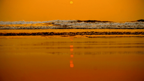 A shot from the surface of the shoreline at a beach in Goa showcasing the twilight view of the sun setting in the horizon and the waves crashing on the shoreline.
